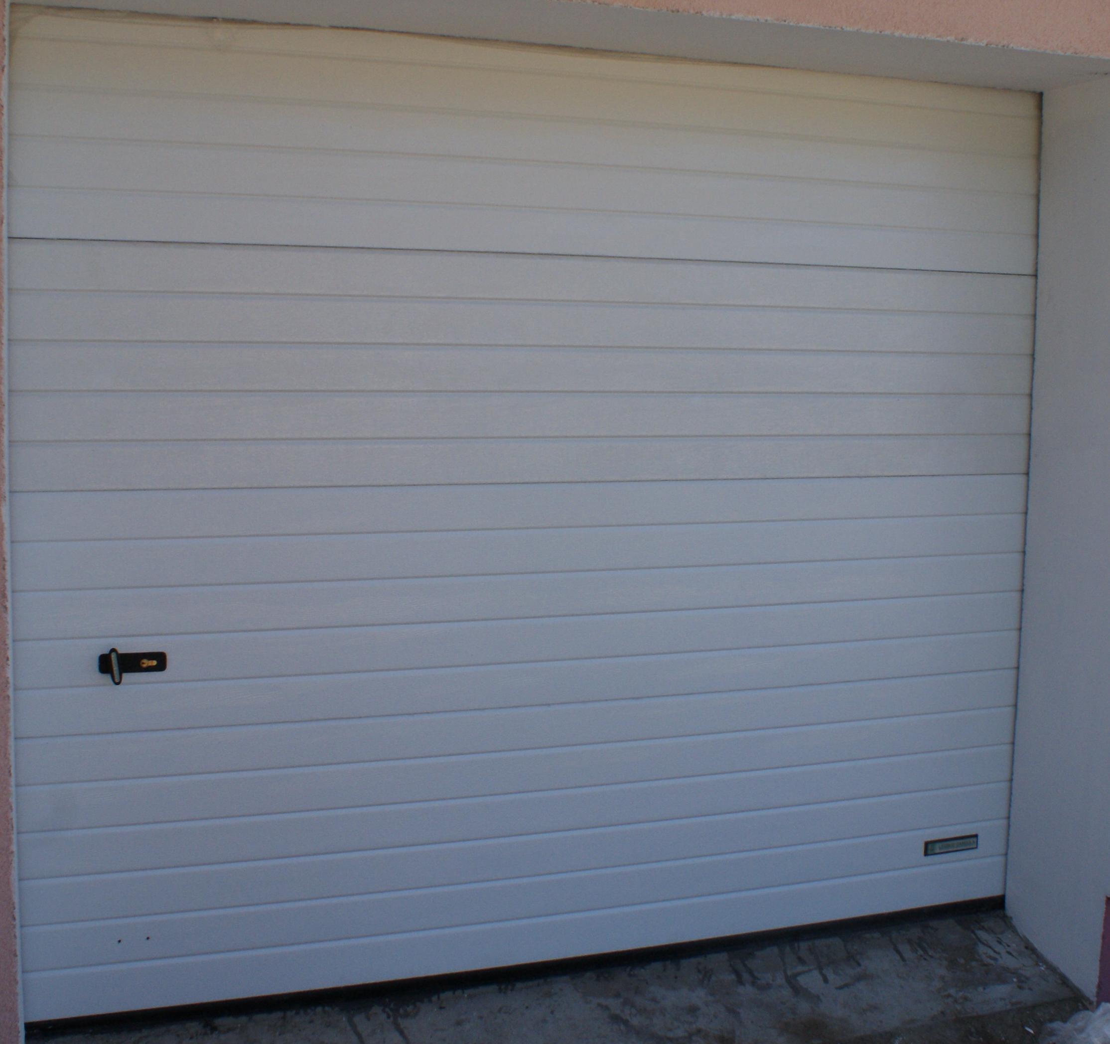 Zamknięta, segmentowa brama garażowa w kolorze białym, z poziomymi przetłoczeniami i czarnym uchwytem, zamontowana w garażu o betonowej posadzce.